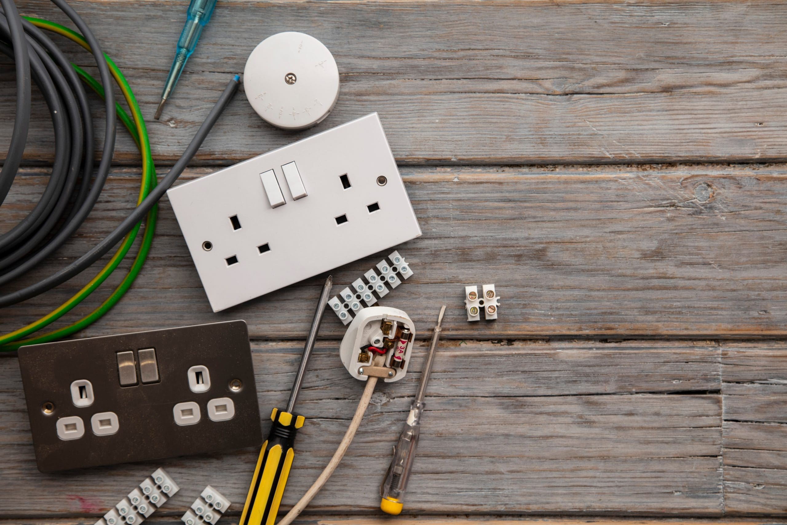 A wooden surface is scattered with various electrical tools and components, including a white double wall socket, black double switch, wires, screwdrivers, and cable connectors—ideal for an electrician conducting rewires or preparing a Landlord Electrical Certificate.