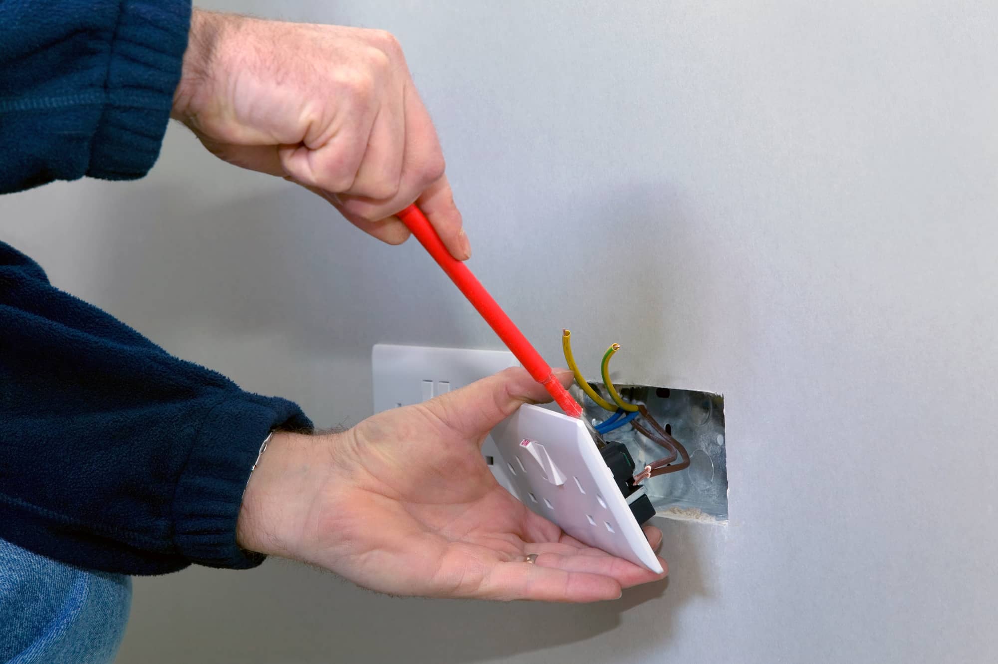 A person uses a red-handled screwdriver to install or remove a wall electrical outlet cover, with exposed wires visible coming from the wall.
