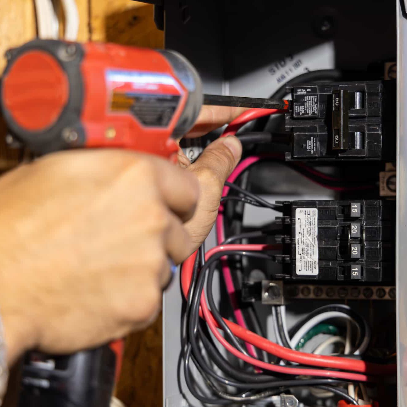 A person uses a red cordless drill to work on an electrical panel filled with wires and circuit breakers.