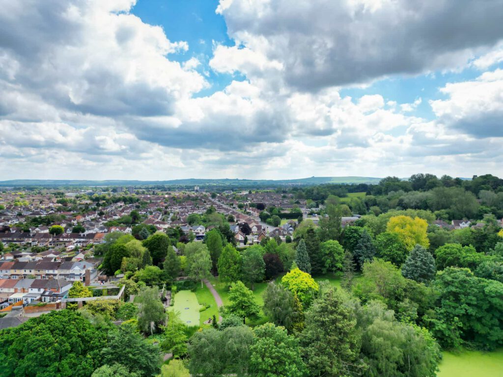 Aerial view of a suburban neighborhood with houses, roads, and lush green trees under a partly cloudy sky, with open fields and hills in the distant background.