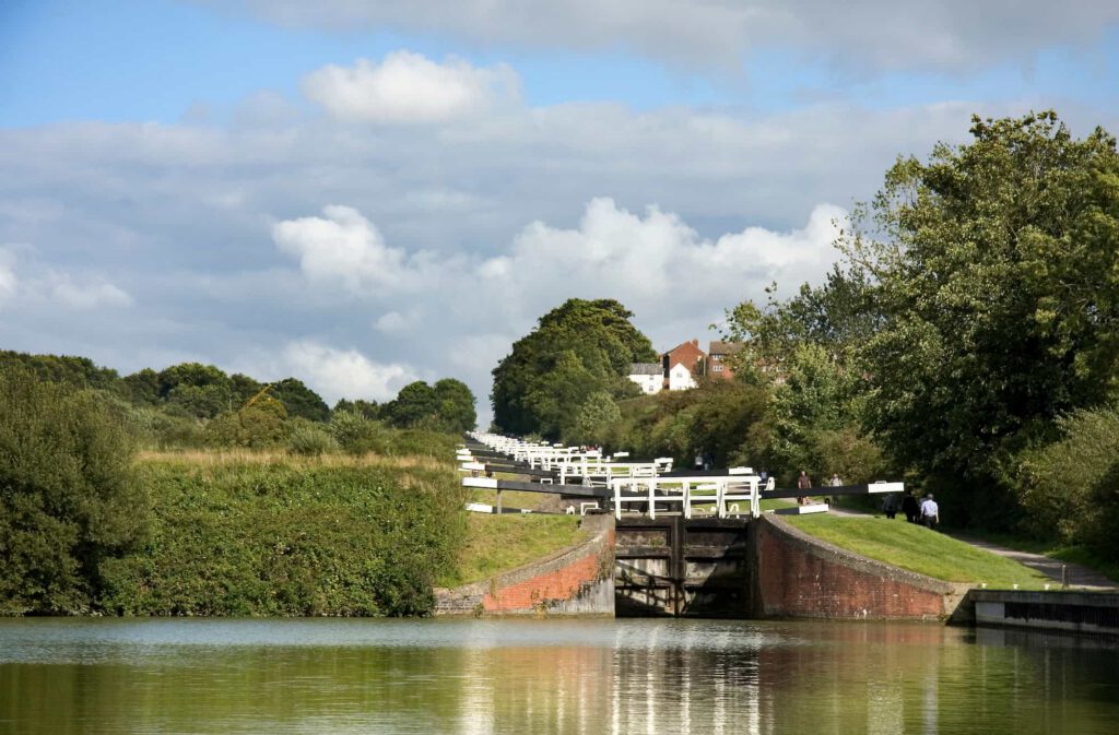 A series of white canal locks stretches up a green hillside beside a calm waterway, with trees and a white house in the background under a partly cloudy sky.