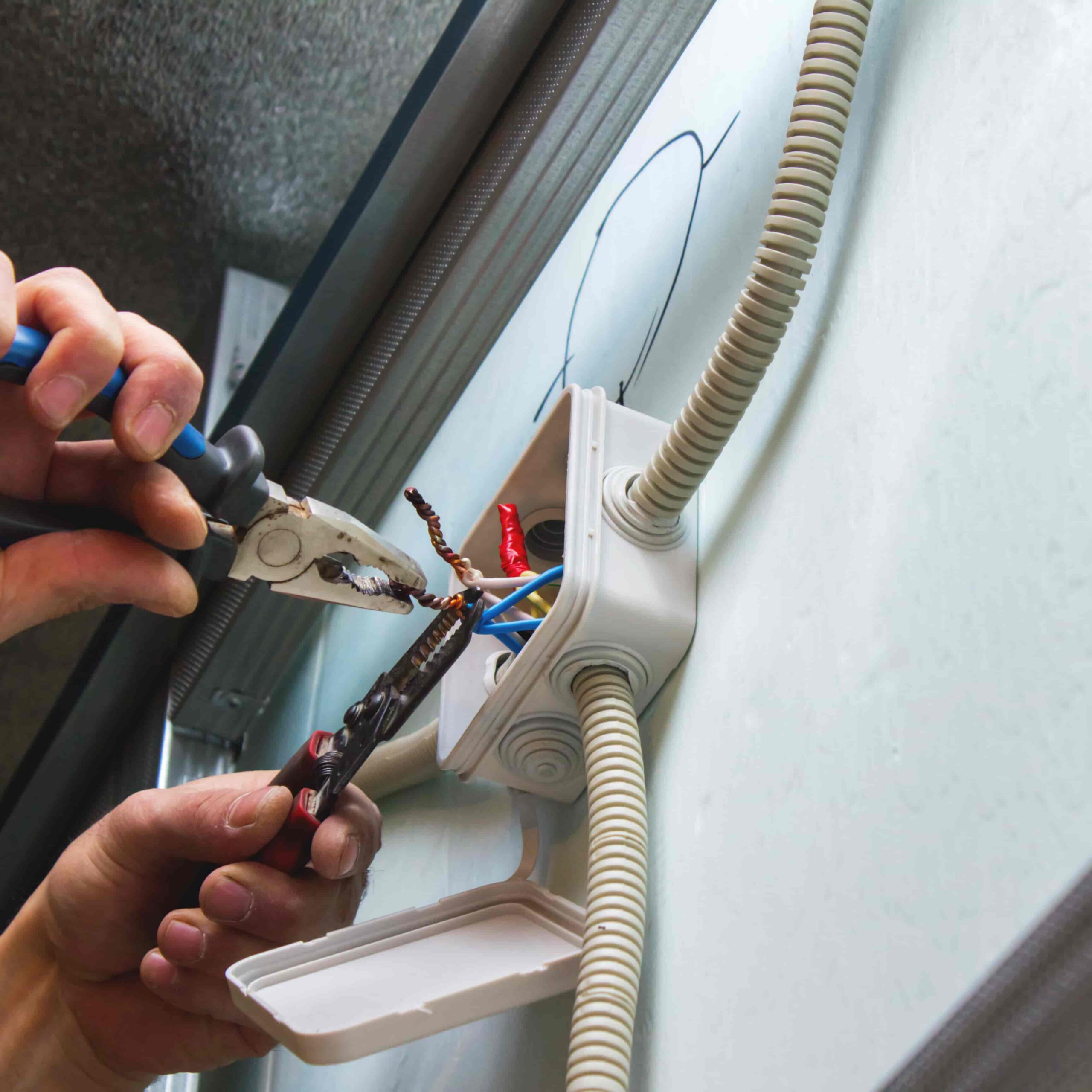 A person uses pliers to twist electrical wires inside an open wall-mounted junction box, with conduit pipes attached to it. The box cover hangs open below.