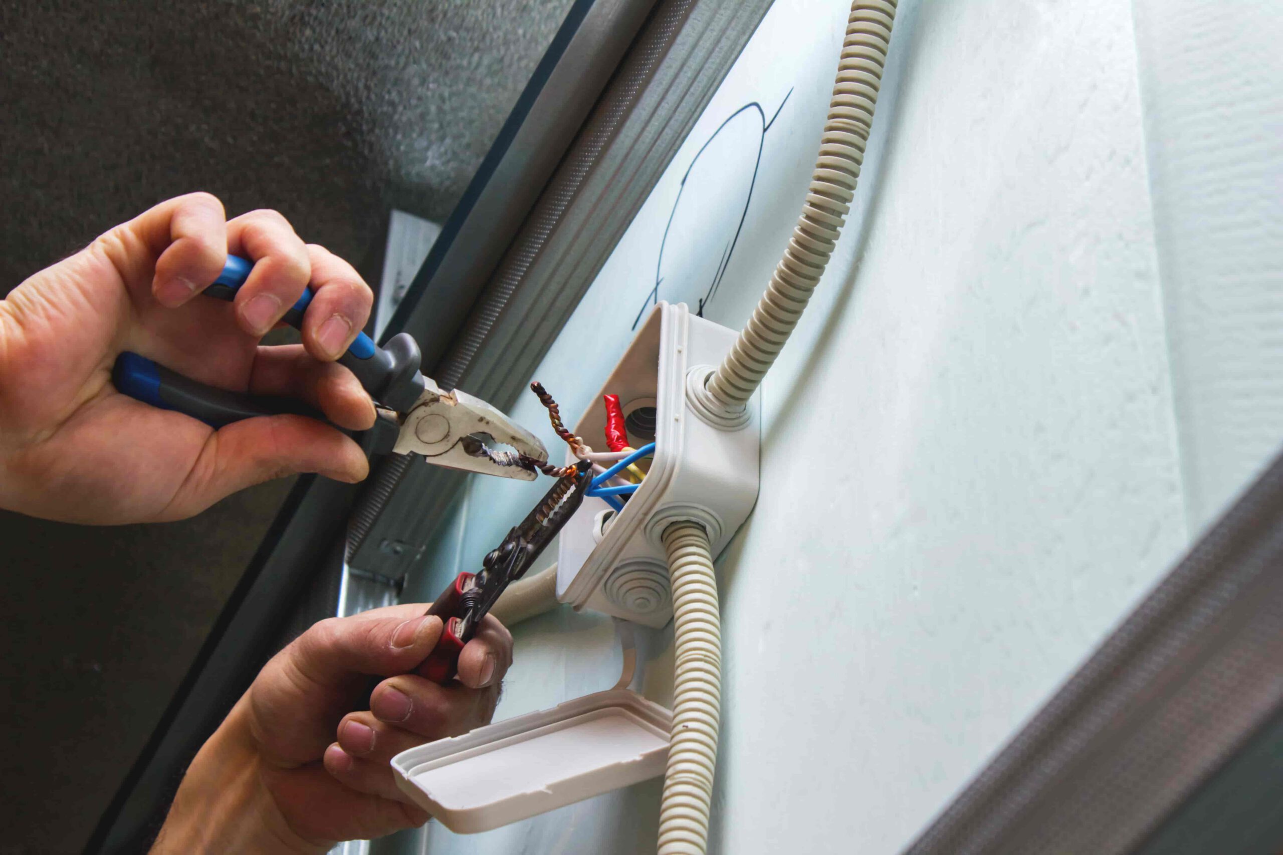 A person uses pliers to work on electrical wires inside a wall-mounted junction box with an open cover, showing several cables and conduits.