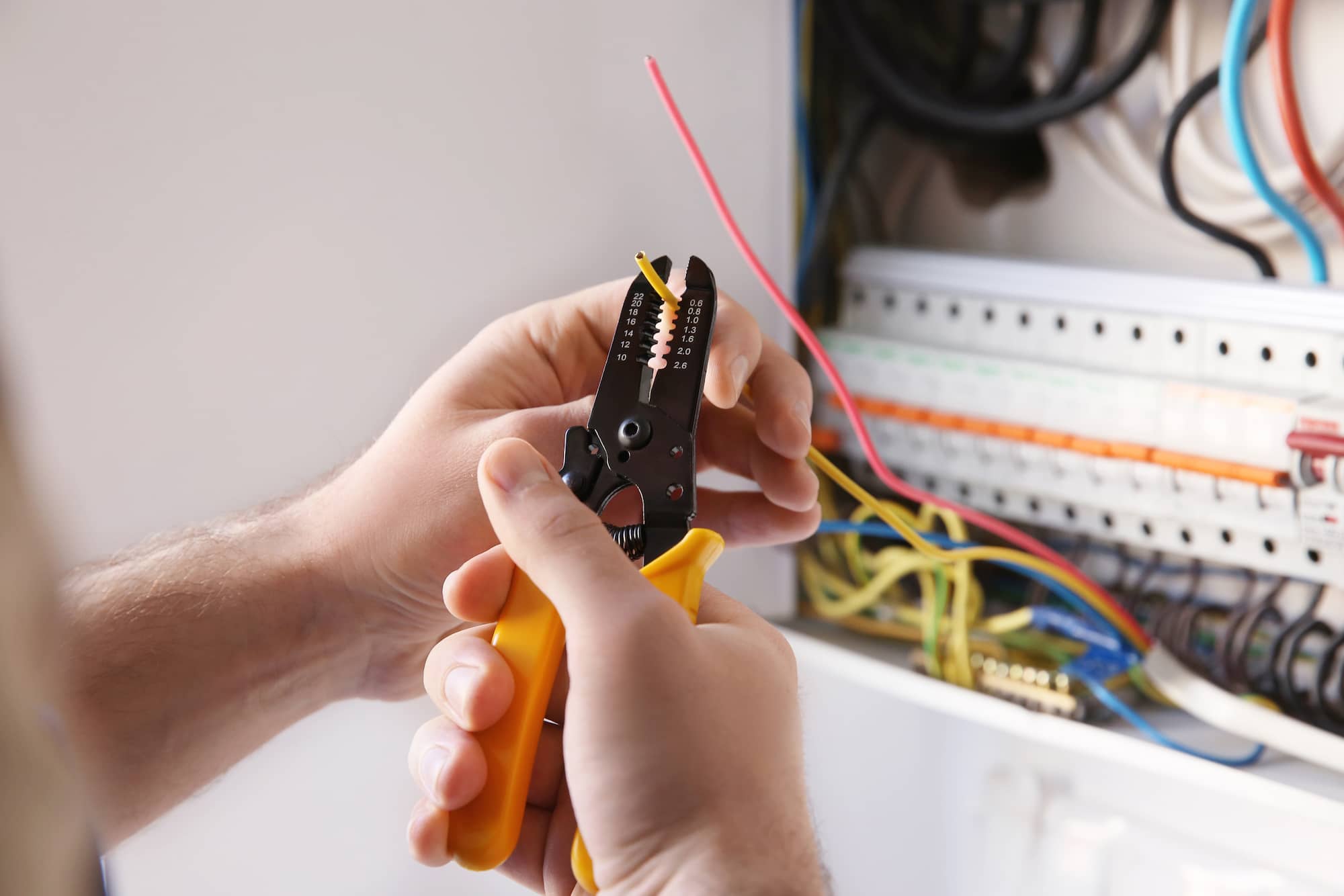 Close-up of hands using a yellow wire stripper to strip insulation from a red electrical wire, with an open electrical panel and various colored wires visible in the background.