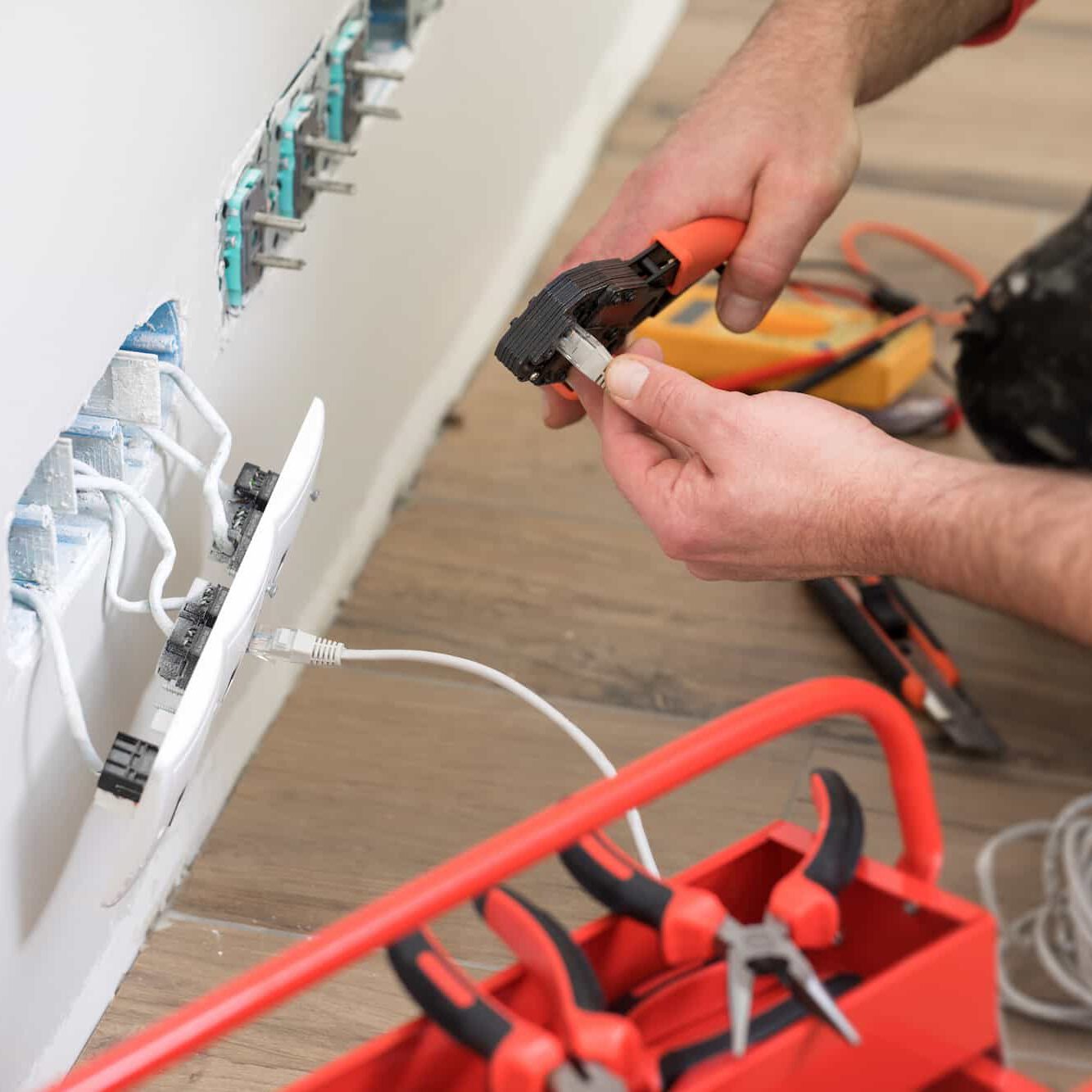 A person uses a crimping tool to attach a connector to a network cable near a wall with open sockets and exposed wiring. A red toolbox with various pliers is visible in the foreground.