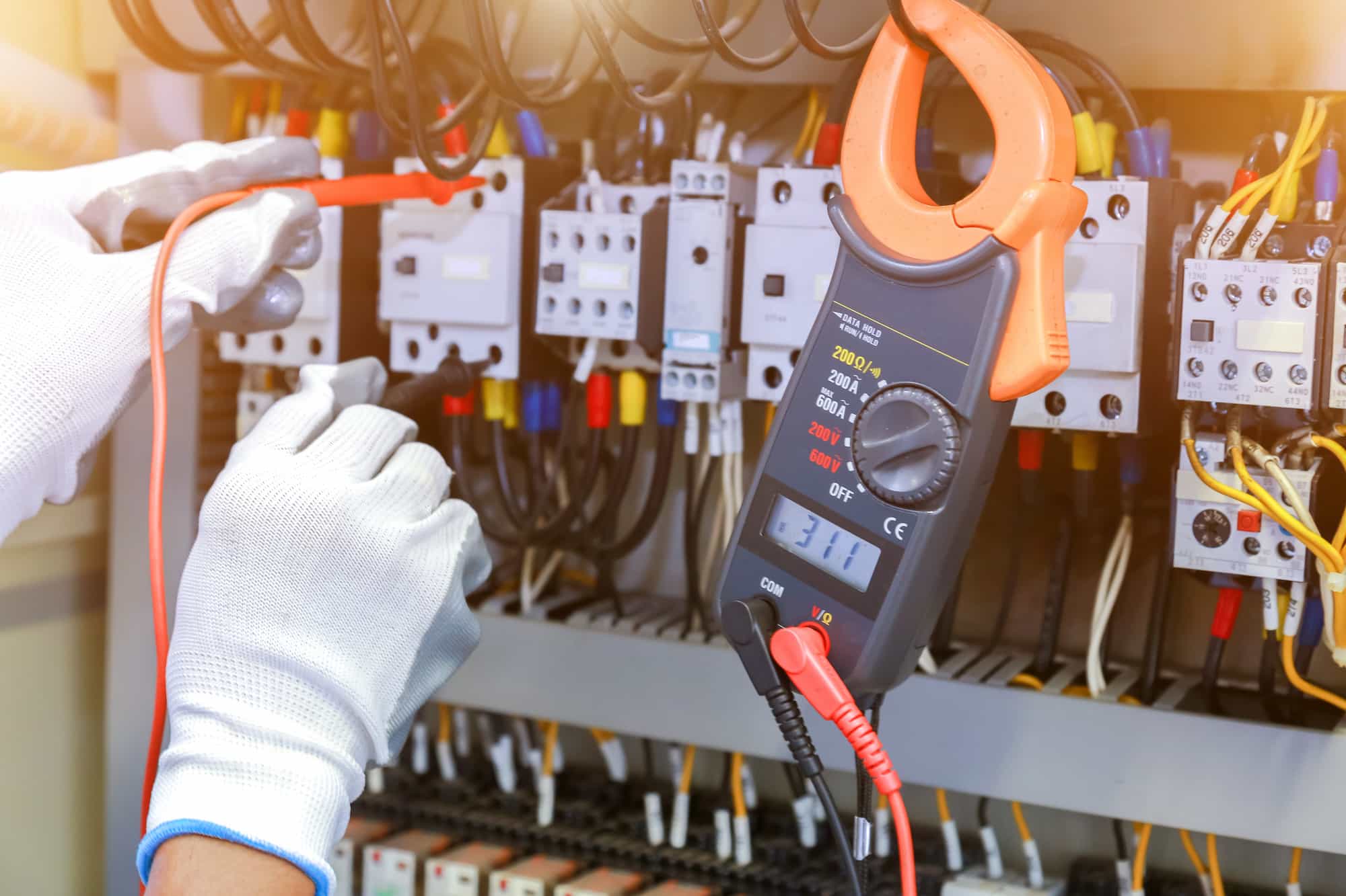 A person wearing white safety gloves uses a digital clamp meter to measure electrical values in an open control panel with multiple wires, switches, and components.