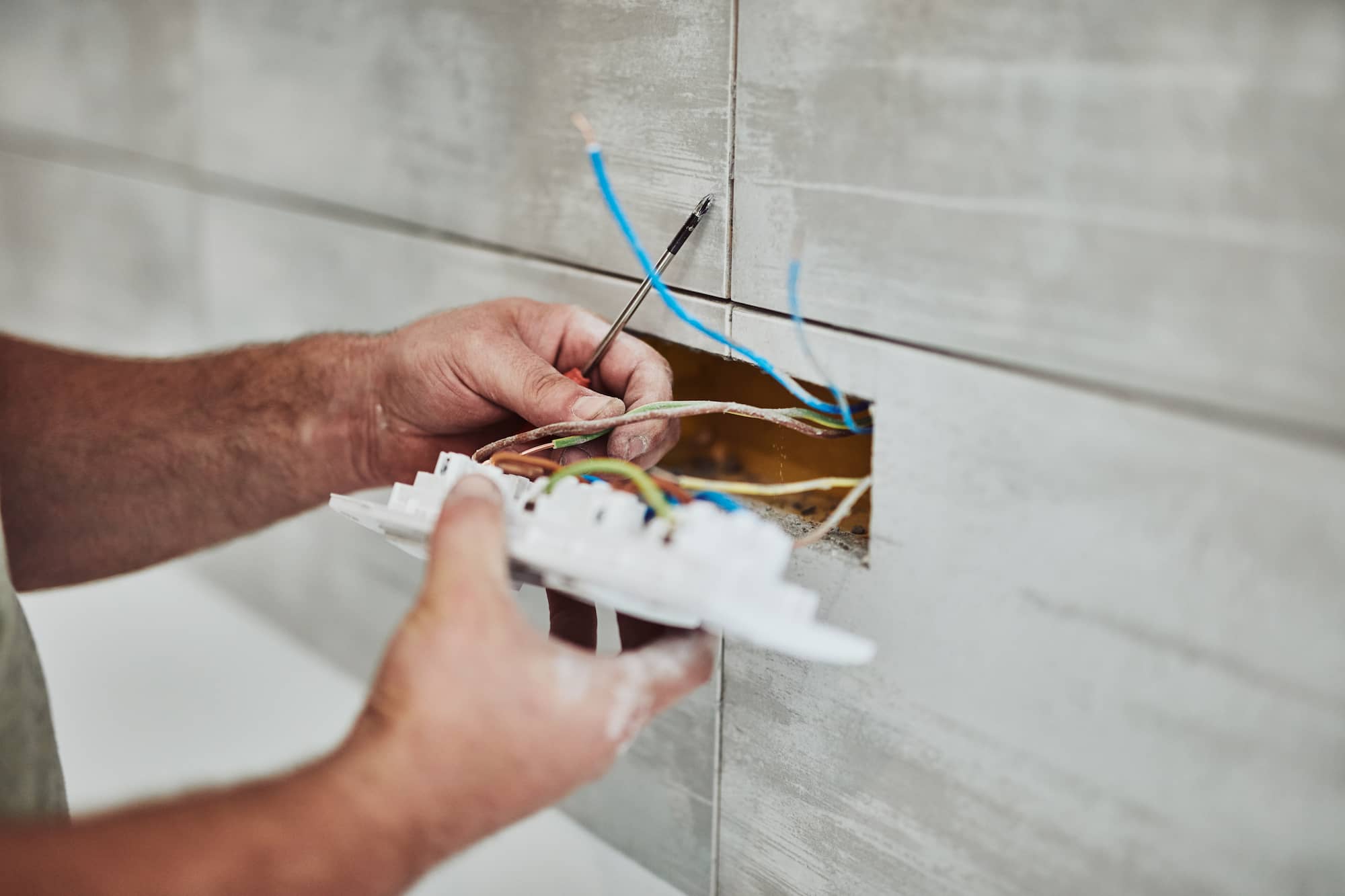 A person installs or repairs electrical wiring in a wall, holding exposed wires and a white electrical fixture near a rectangular hole in a tiled wall.