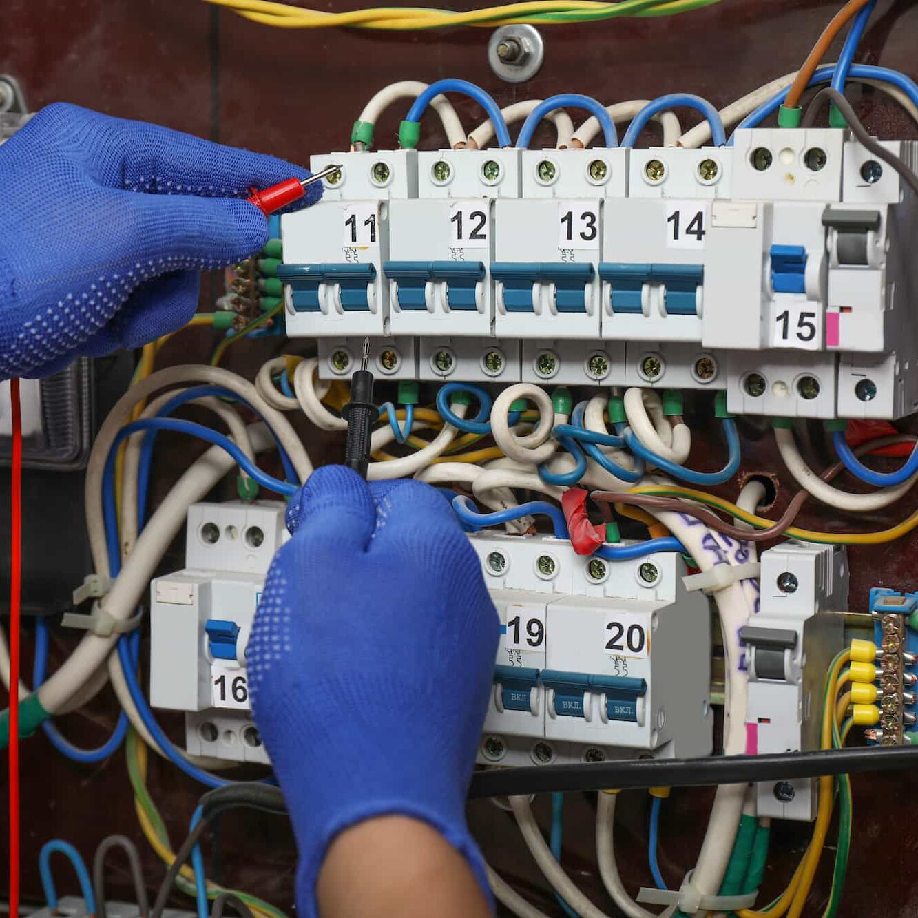 A person wearing blue gloves uses probes to test electrical circuit breakers on a panel filled with numbered switches and a tangle of colored wires.
