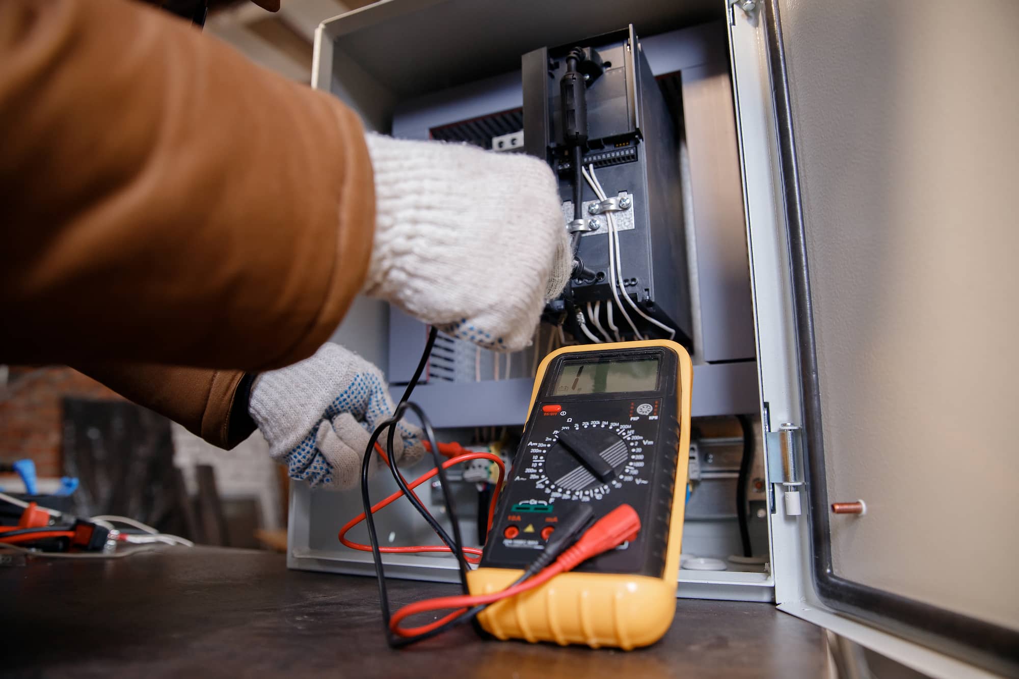 A person wearing gloves uses a multimeter to test electrical connections inside an open control panel. The multimeter is in the foreground, with wires and components visible inside the panel.