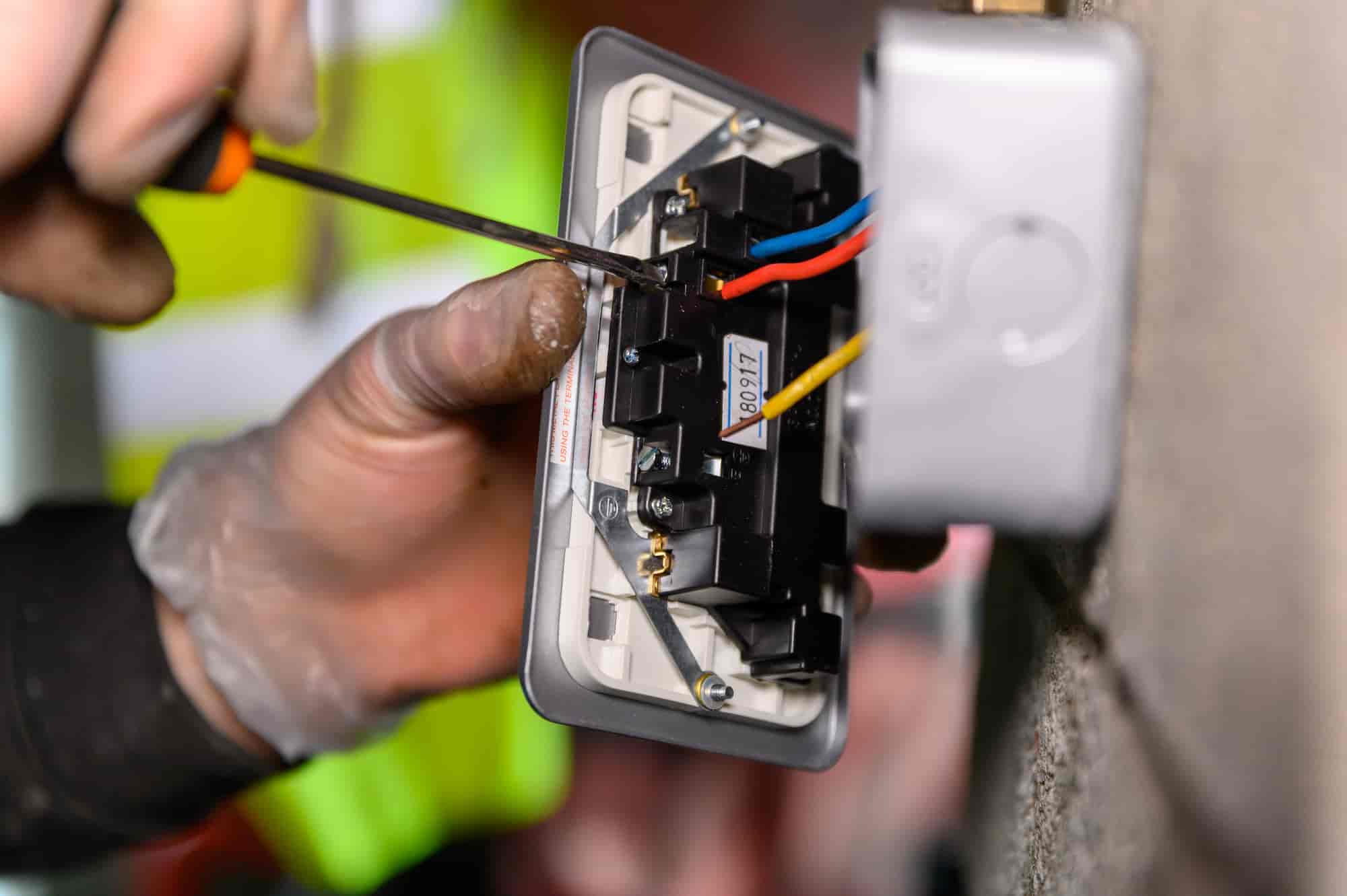 A close-up of a person’s hand using a screwdriver to wire an electrical socket, with red, blue, and yellow wires visible. The person is wearing a glove and a high-visibility jacket can be seen in the background.