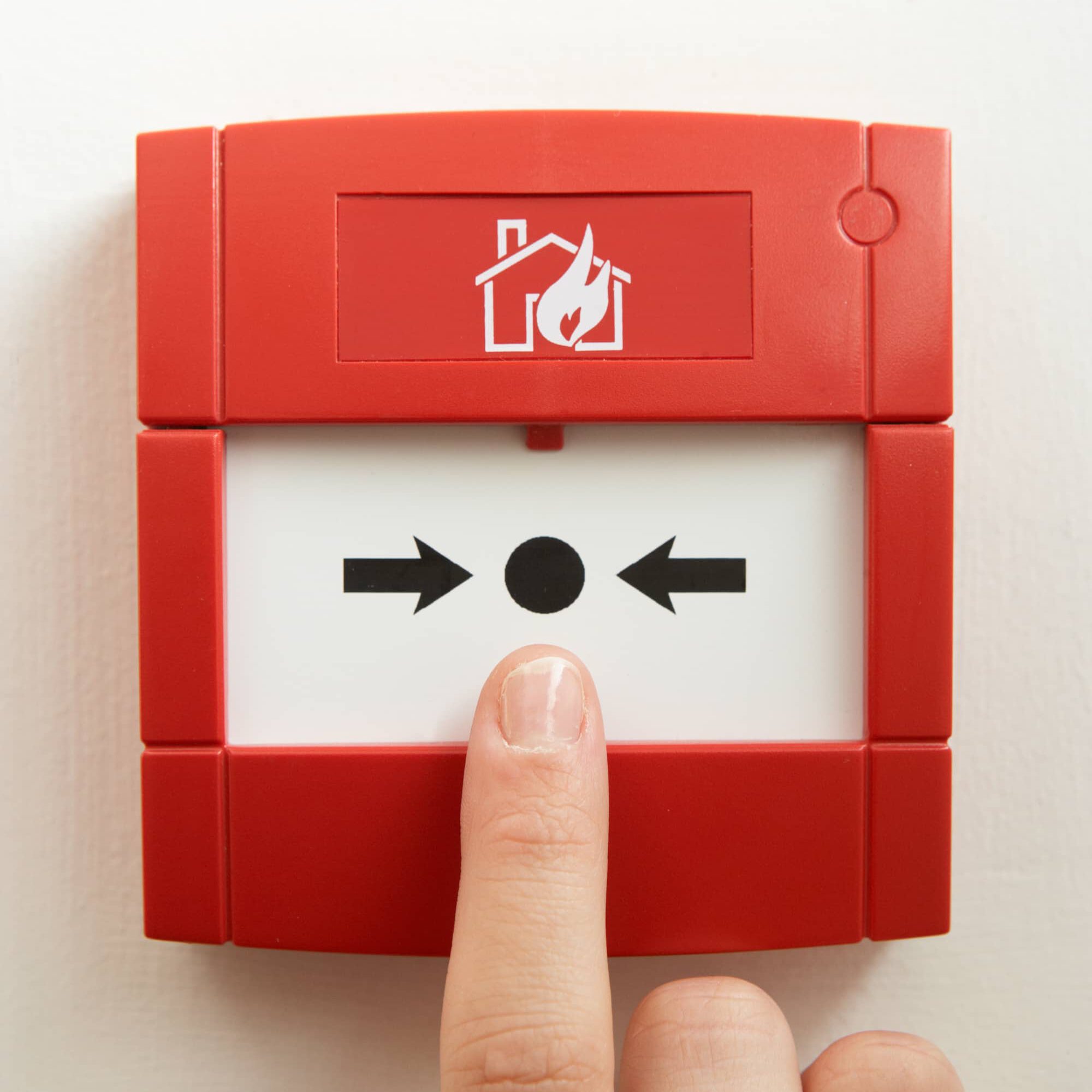A finger pressing the center button on a red fire alarm call point mounted on a white wall. The device has a house with flames icon, and black arrows pointing toward a black dot in the middle.