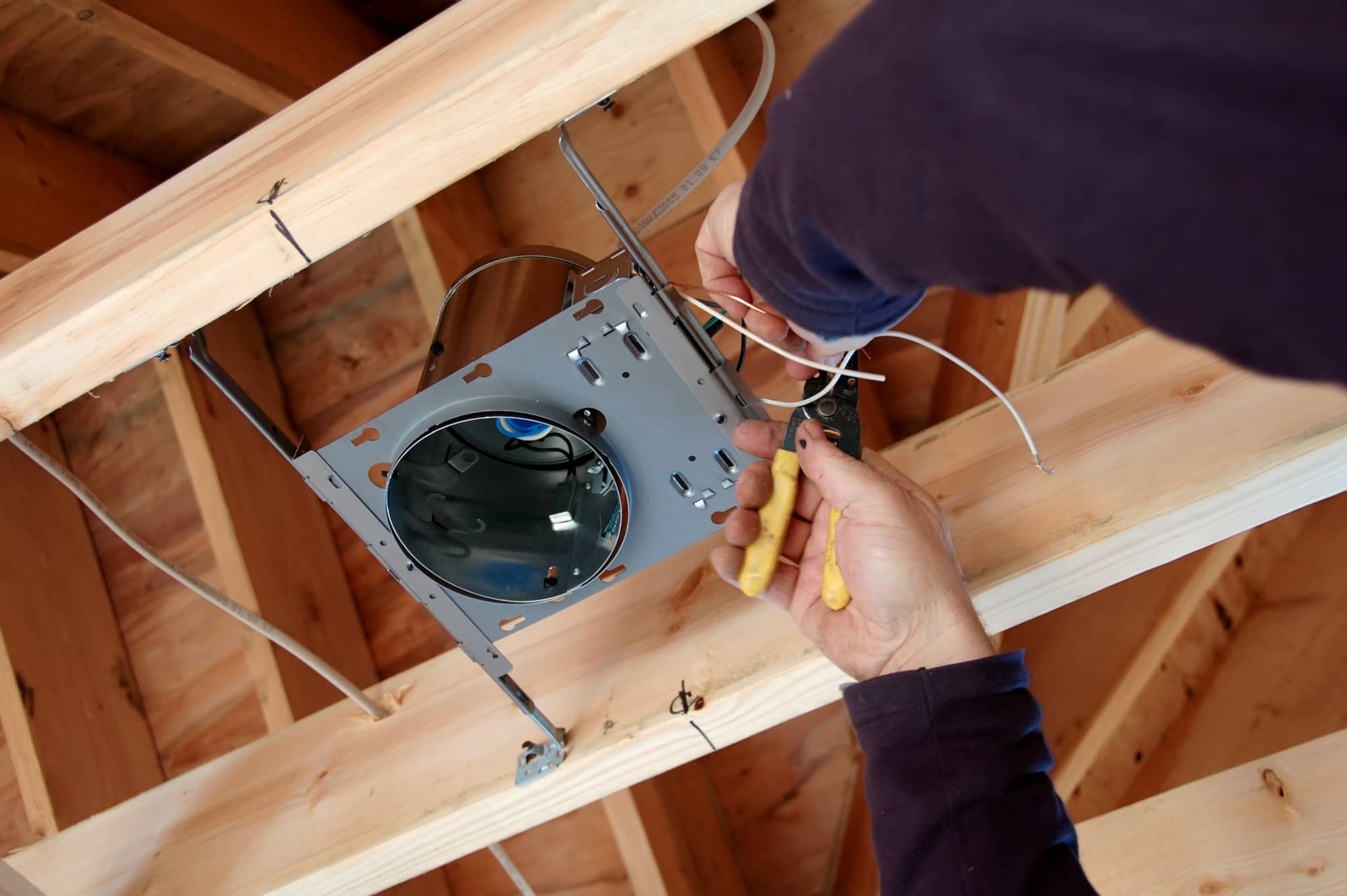 A person installs electrical wiring for a recessed light fixture in a wooden ceiling, using wire cutters and preparing connections.