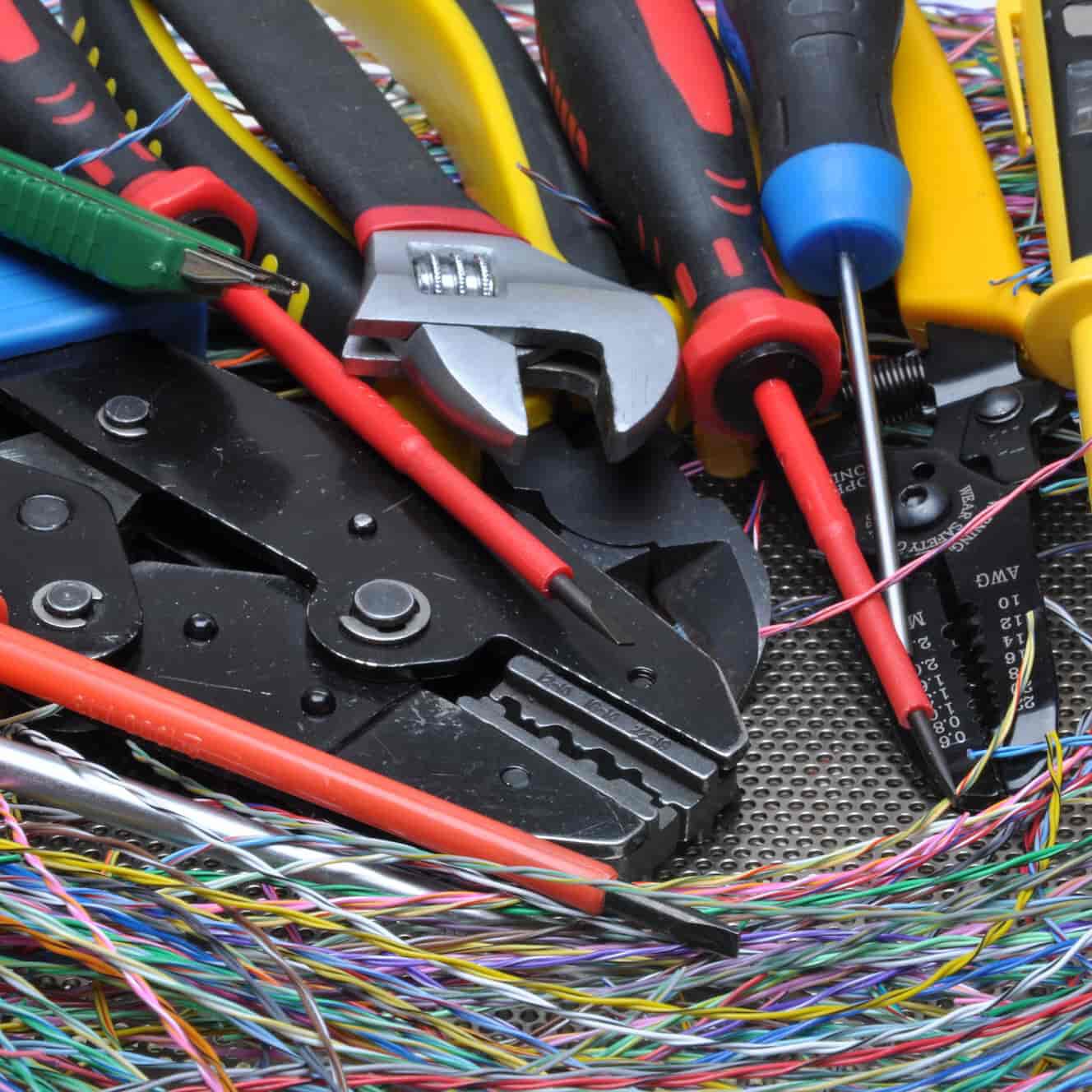 A close-up of various hand tools, including pliers, screwdrivers, a wrench, and wire strippers, surrounded by colorful electrical wires.