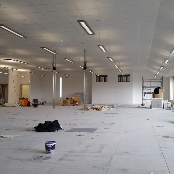 A large, empty room under construction with scattered building materials, a paint bucket, black cloth, and ladders. Ceiling lights are installed, and the floor is unfinished with visible construction marks.