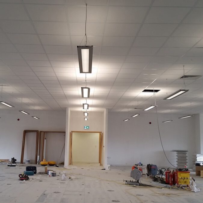 An unfinished office space with white walls, exposed ceiling lights, construction tools, and equipment scattered on the floor; some ceiling tiles and light fixtures are missing.